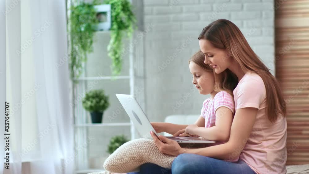 Positive blonde female and pretty child use computer sitting on bed. Medium shot on RED camera Positive blonde female and pretty child use computer sitting on bed. Medium shot on RED camera