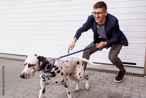 Photography A young man having troubles holding a dalmatian dog on a leash, on the white roller shutters background