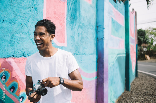 Laughing Hispanic man in casual clothes with camera on street