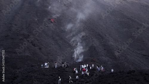 A big group of tourists stand dangerously close to a smoking lava flow as a large molten rock rolls down the black volcano towards them photo