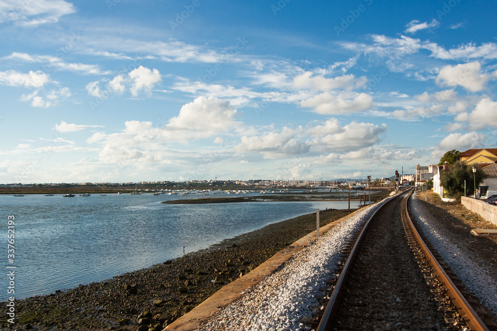 Railway by the shore. Atlantic ocean in Faro Portugal. Sunset landscape. Calm water bay with boats floating. Travel by train around the Europe - cloudy sky panoramic view.