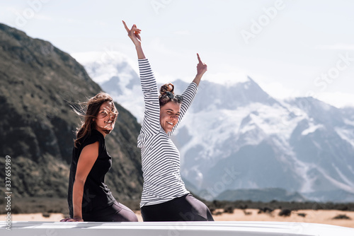 Two happy young girls who are travel lovers enjoy the moment side by side on a roadtrip. They are on top of the van laughing in front of the mountains and getting lost in the nature.Travel concept.