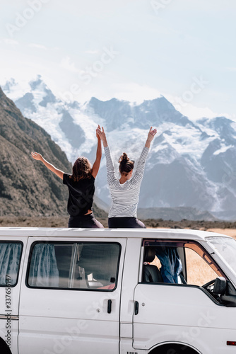 Two adventurous female travelers raise their hands in front of Mount Cook lookout. They pull over to the side and sit back on top of the van to enjoy the moment together. Copyspace. Travel concept