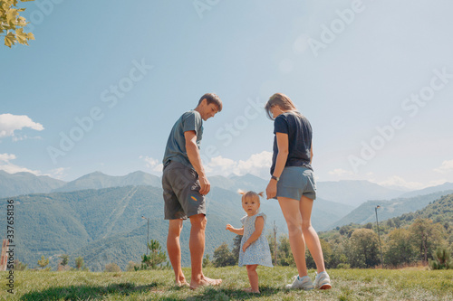 Beautiful young family, parents and little daughter, looking at the camera,  standing  on the green lawn with great mountain view backwards in the sunny day, blue sky
