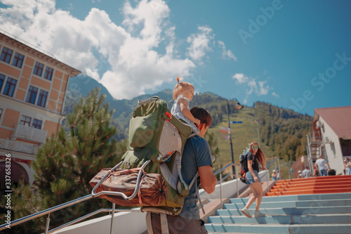 Father and beautiful little daughter on his back, and her mother looking at them near, walking on the colorful stair in the sunny day, great mountains view, interesting buildings backwards
