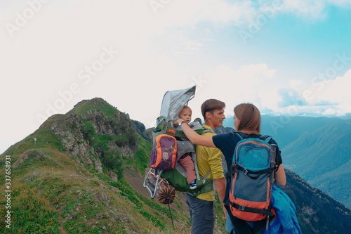 Young beautiful family, parents and child in the child carrier, hiking on the picturesque mountain trail in the summer forest with great mountain view with blue sky backwards, sunny day 