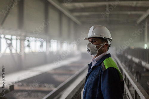 Mining engineer in yellow-blue uniform and white helmet and respirator inspects dusty and dirty mining workshop. Selective focus © Model Republic