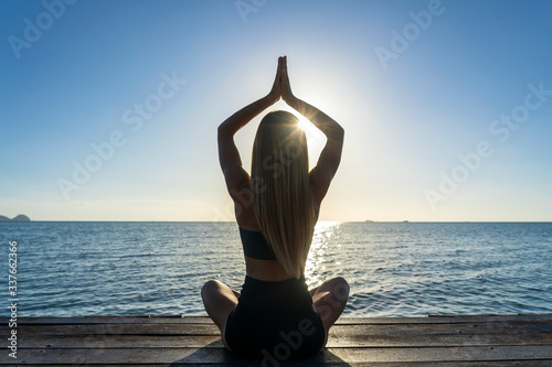 Silhouette of woman sitting at yoga pose on the tropical beach during sunset. Girl practicing yoga near sea water
