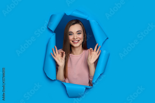 Portrait of beautiful joyful smiling caucasian woman showing Ok gesture and posing in pink paper hole. copy space.