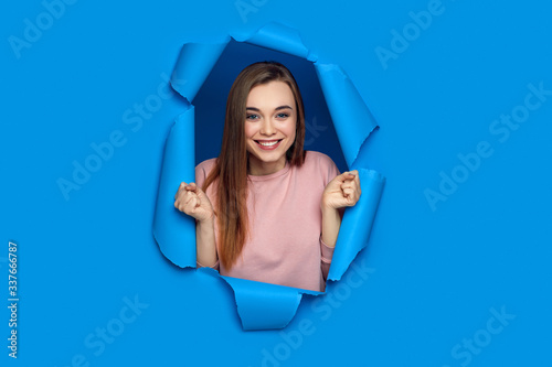 Portrait of beautiful happy smiling caucasian woman raises clenched fists and poses in blue paper hole. © producer