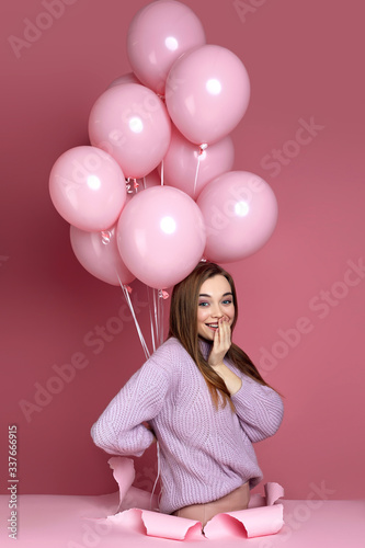 surprised cute woman with pastel pink air balloons standing in torn paper hole on pink background. Beautiful happy young girl as a gift for birthday holiday.