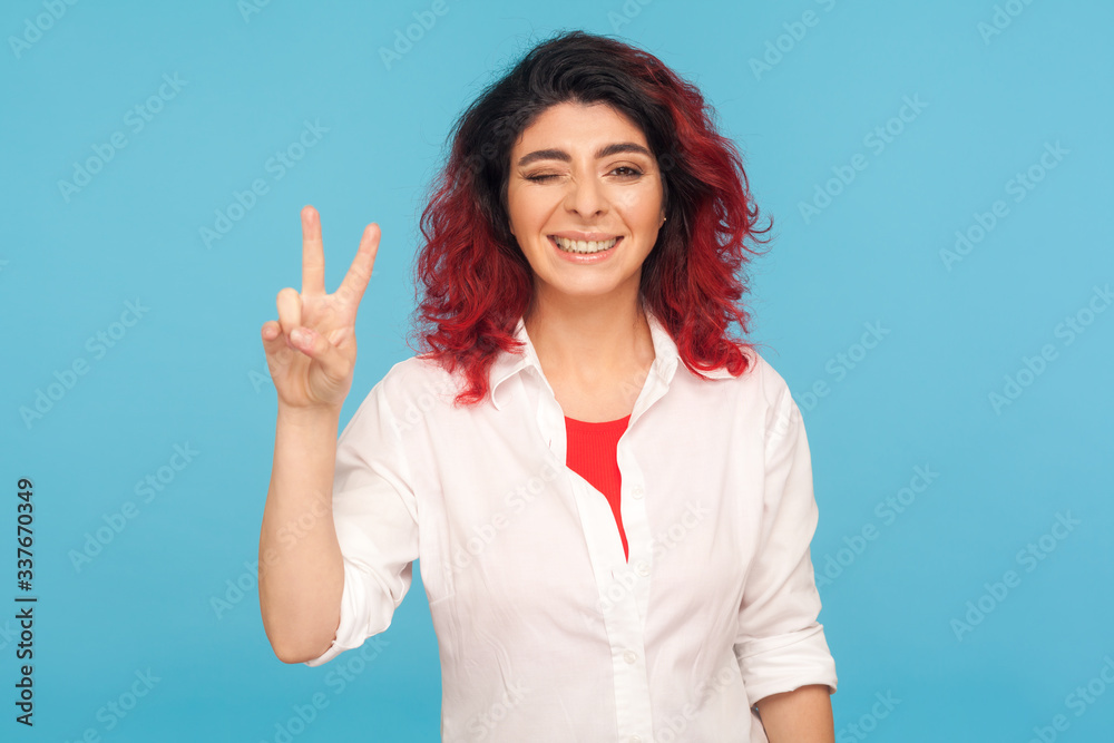 Portrait of happy successful hipster woman with fancy red hair winking playfully to camera and showing victory gesture, making peace sign with fingers. indoor studio shot isolated on blue background Portrait of happy successful hipster woman with fancy red hair winking playfully to camera and showing victory gesture, making peace sign with fingers. indoor studio shot isolated on blue background