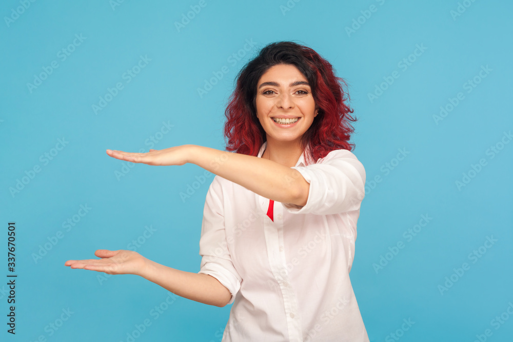 Awesome hipster woman with fancy red hair presenting advertising area between hands, holding copy space for commercial and smiling pleased satisfied. indoor studio shot isolated on blue background Awesome hipster woman with fancy red hair presenting advertising area between hands, holding copy space for commercial and smiling pleased satisfied. indoor studio shot isolated on blue background