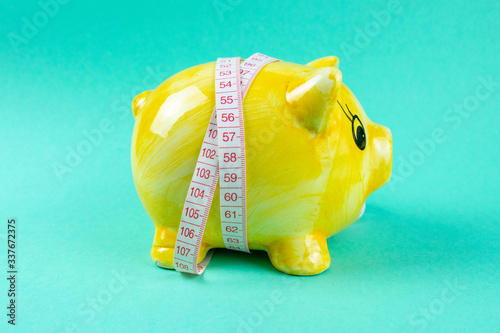 Yellow piggy bank in the form of a pig wrapped with measuring tape on a light green background. The concept of saving money for the sake of losing weight