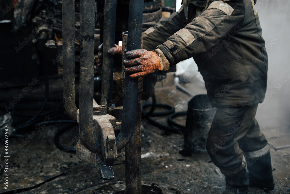 Work driller in red uniform, in helmet and goggles. He uses a hydraulic wrench to screw drill pipes to lower them into an oil well and continue drilling it. The concept of a working person. Work driller in red uniform, in helmet and goggles. He uses a hydraulic wrench to screw drill pipes to lower them into an oil well and continue drilling it. The concept of a working person.