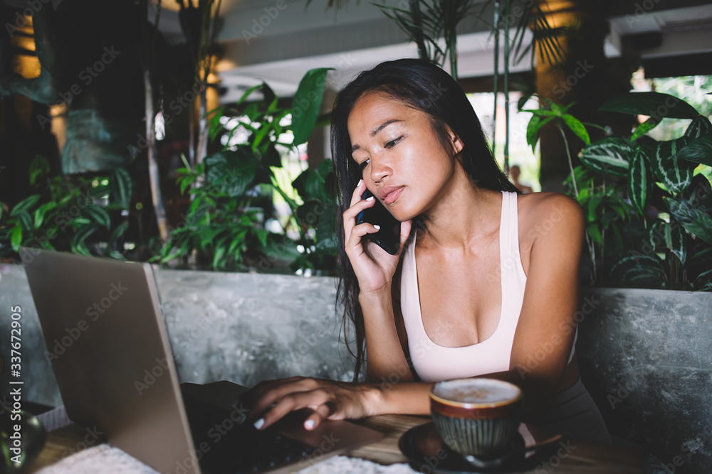 Young Asian woman typing on laptop while making call