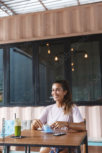 Vertical portrait of pretty european woman sit at cafe table with smoothie and mobile phone, drinking coffee, laughing and enjoying perfect vacation on relaxing resort Bali island, smiling pleased © Liubov Levytska
