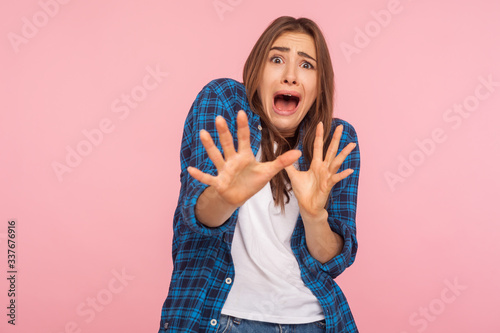 I'm afraid! Portrait of shocked scared girl in checkered shirt screaming in horror and fear, keeping hands raised to defend herself, panic attack. indoor studio shot isolated on pink background