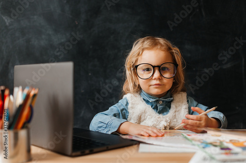 pretty little girl in glasses sits at her desk and learns online, looking at the camera with a smile