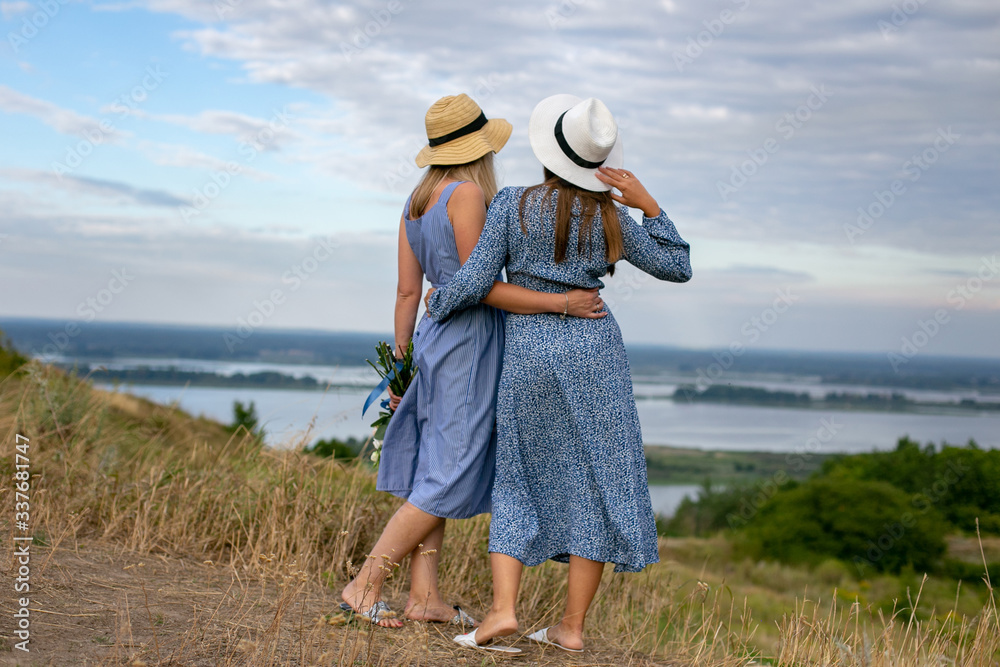 horizontal portrait of two long-haired European women in summer dresses and hats against the backdrop of beautiful nature