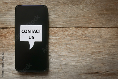 Top view of a mobile phone with a white speech bubbles written with Contact Us on wooden background.