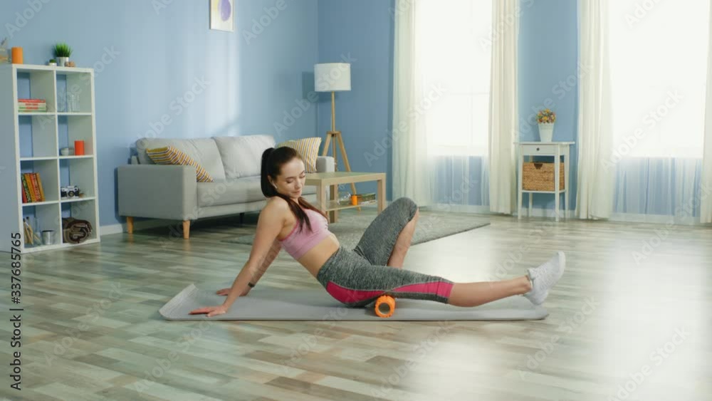 Young pretty woman is workouting at home, using foam roller to massage deep tissue, enjoying her morning training, listening favorite music, relaxing, slow motion. Young pretty woman is workouting at home, using foam roller to massage deep tissue, enjoying her morning training, listening favorite music, relaxing, slow motion.