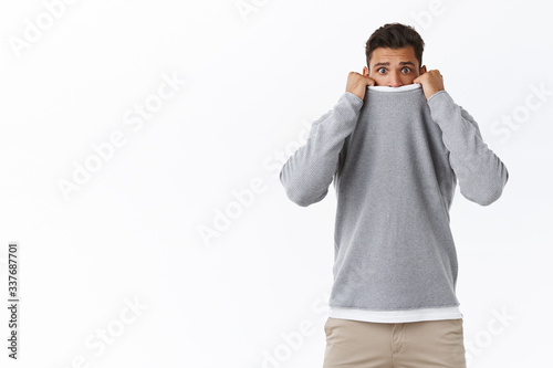 Scared, timid and cute young guy feeling afraid of horror movies, pulling sweater on face and frowning with arched sad expression, being frightened, standing shaking fear white background