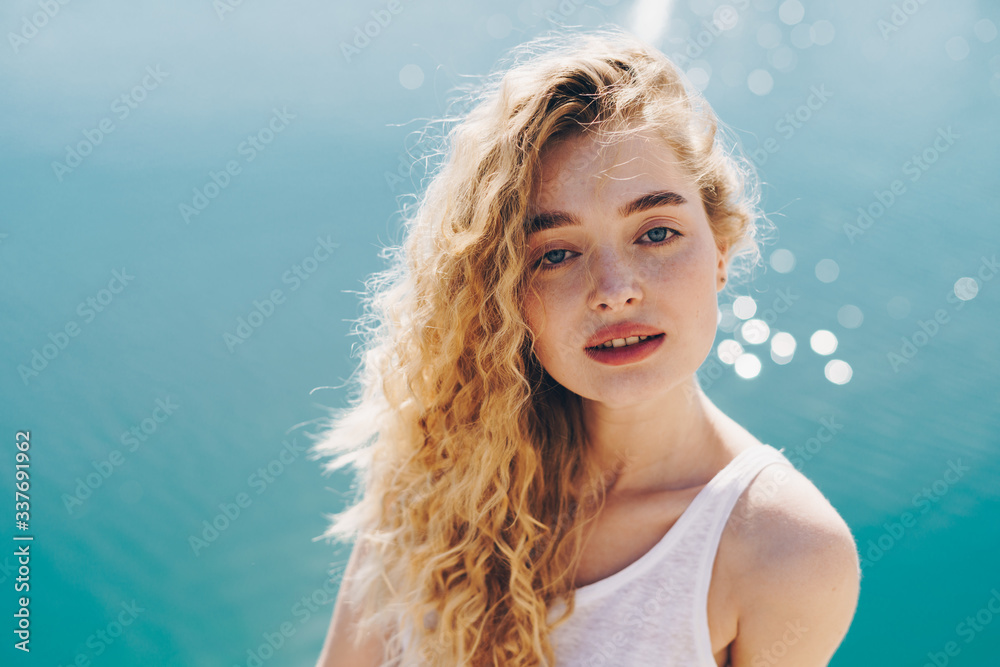 Mysterious curly blonde on a background of blue sea looks at the camera. Mysterious curly blonde on a background of blue sea looks at the camera.