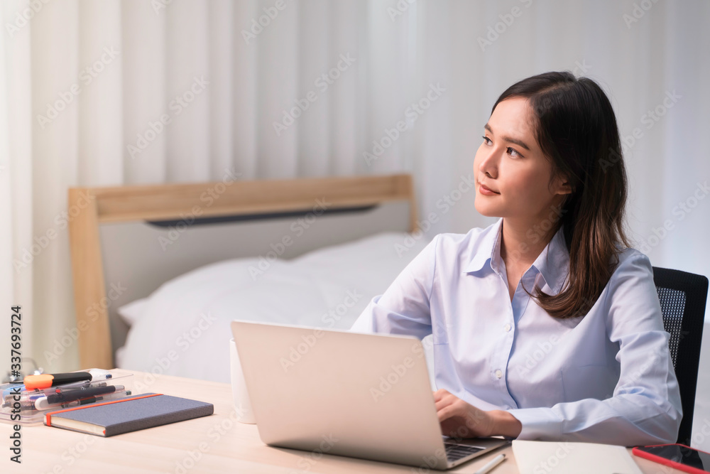 Attractive Asian woman works from home with laptop on desk in bedroom. Young girl smiling with happy face, looking outside and thinking about work. Shelter in place from COVID-19 and Stay Home concept Attractive Asian woman works from home with laptop on desk in bedroom. Young girl smiling with happy face, looking outside and thinking about work. Shelter in place from COVID-19 and Stay Home concept