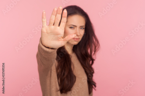 No, denial concept. Portrait of young woman with brunette wavy hair standing with outstretched hand making stop gesture, prohibition or rejection sign, warning of danger. studio shot, pink background photo