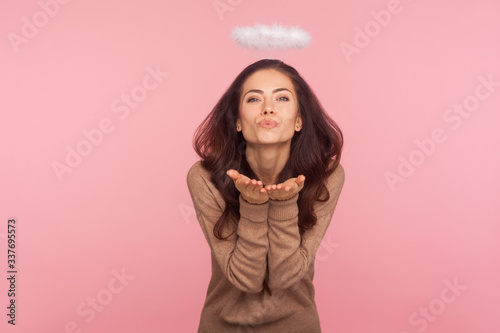 Portrait of beautiful sexy young woman with halo above head sending sensual air kiss expressing love feelings, seducing with angelic beauty, flirting. indoor studio shot isolated on pink background © khosrork