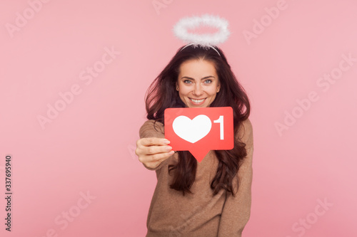 Social media Like button. Portrait of happy angelic young woman with halo over head holding internet Heart icon, recommending to follow and love content. indoor studio shot isolated on pink background