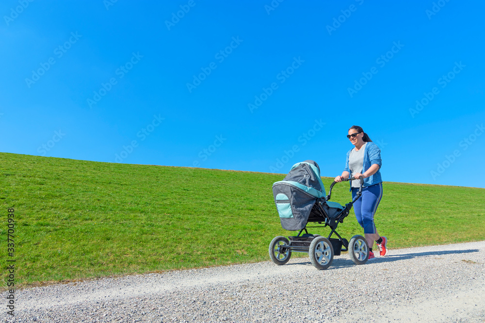 Smiling young mother pushing baby stroller. Green grass and blue sky background Smiling young mother pushing baby stroller. Green grass and blue sky background