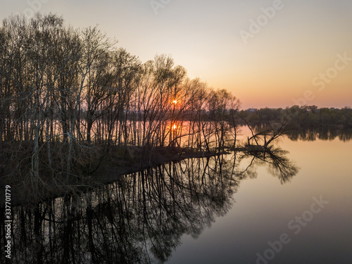 The rays of the setting sun through rare trees on the island in the lake. Aerial drone view.