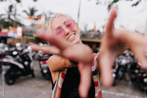 Adorable young woman in pink sunglasses expressing positive emotions on blur street background. Outdoor shot of graceful tanned girl waving hands in front of camera.