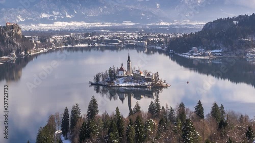 Time lapse zoom in view of Lake Bled island church and boats in winter landscape. Calm lake water reflects sky clouds above, snow covered mountains in the background
