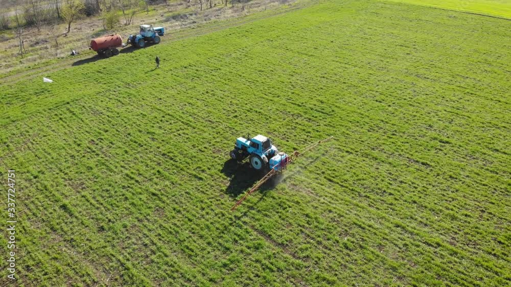 Tractor with mounted sprayer with liquid chemicals process green field of barley or wheat with working people and trailer tank. Spring agronomic activity Tractor with mounted sprayer with liquid chemicals process green field of barley or wheat with working people and trailer tank. Spring agronomic activity