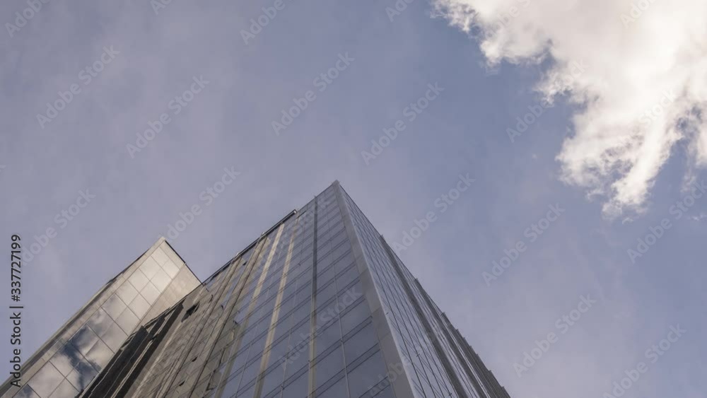 Zoom out time lapse skyscraper with clouds moving above in sky and reflected on the glass outer walls, looking up angle view Zoom out time lapse skyscraper with clouds moving above in sky and reflected on the glass outer walls, looking up angle view