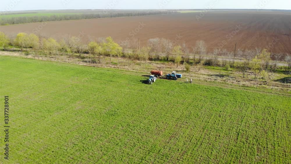 Aerial drone shot two farmer tractors with trailer tank and mounted sprayer with liquid fertilizer on green crop field with working people. Spring agronomic activity Aerial drone shot two farmer tractors with trailer tank and mounted sprayer with liquid fertilizer on green crop field with working people. Spring agronomic activity