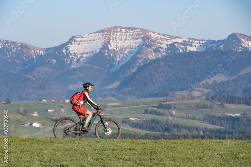 pretty senior woman riding her electric mountain bike in early springtime in the Allgau mountains near Oberstaufen, in warm evening light below the spectacular snow capped mountains of Nagelfluh chain