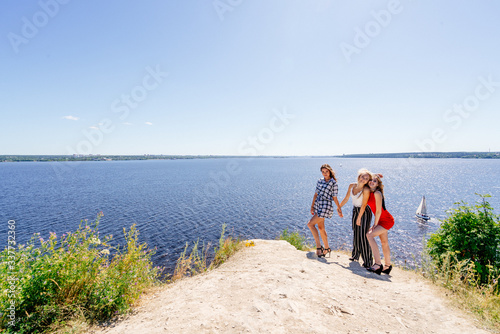 Shoot of three beautiful girls outdoors by the river. Female friends relaxing by the river and smiling. Girlfriends are stand on the pier, joy, fun, hands up. Girls with drinks in dresses. Summer