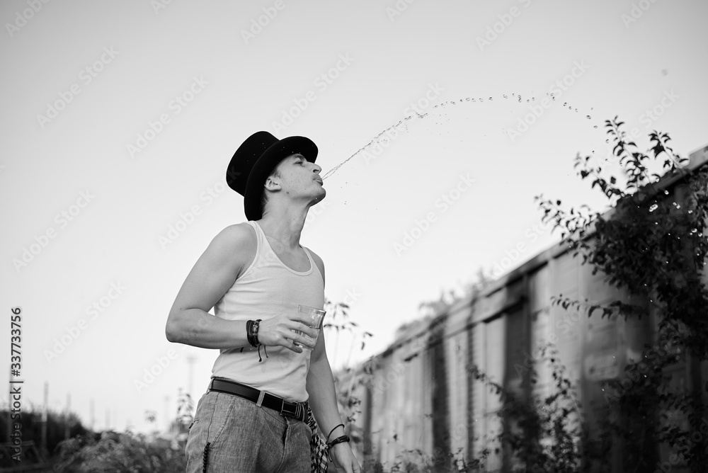 Φωτογραφία Three-quarter portrait of young man, wearing grey pants,white top, black classic hat, holding water glass, spitting on the side