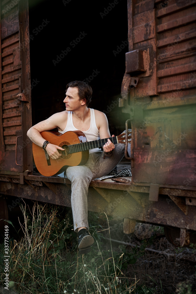 Fototapeta premium Young handsome man, wearing white t-shirt and grey pants, sitting inside old railway carriage, playing guitar, composing music Brunette male musician singer with guitar on abandoned train in summer.