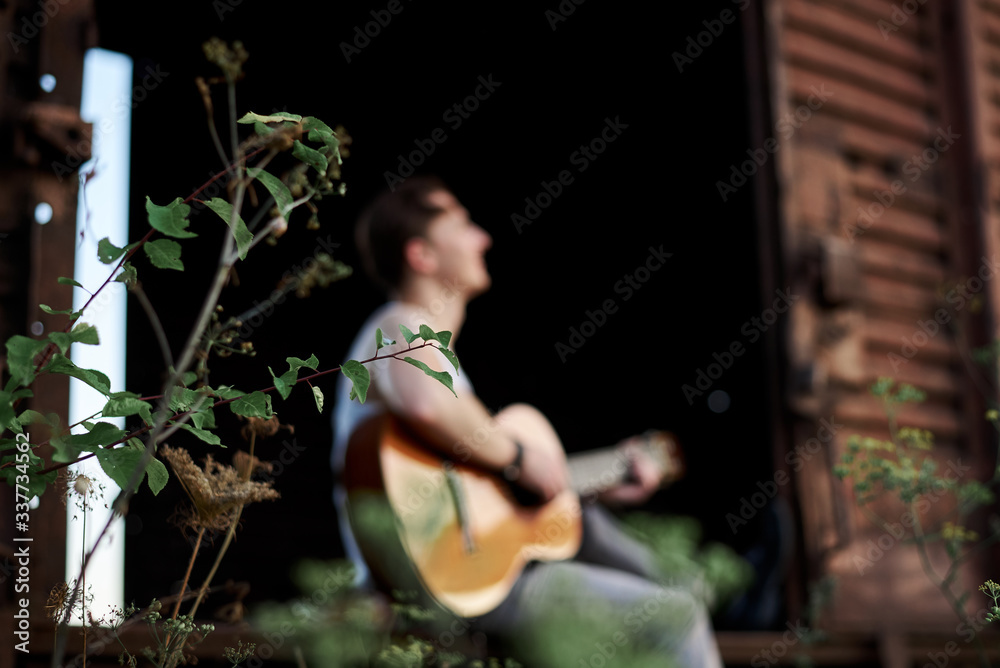 Young handsome man, wearing white t-shirt and grey pants, sitting inside old railway carriage, playing guitar, composing music Brunette male musician singer with guitar on abandoned train in summer.