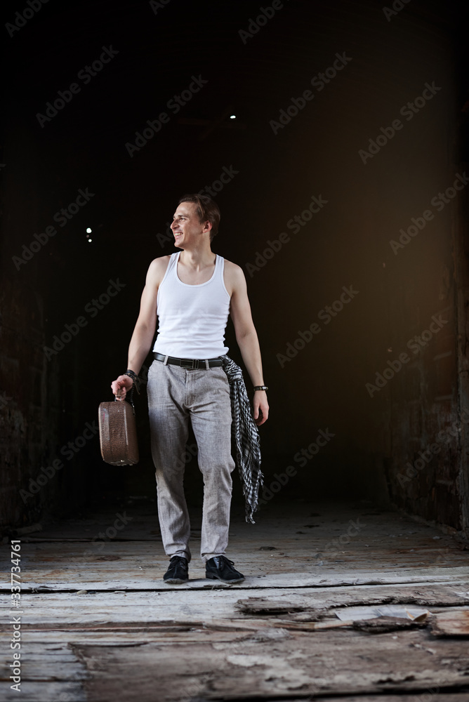 Full-length portrait of young man, wearing white t-shirt, grey pants and black magician hat, walking inside old railway carriage holding old suitcase. Creative man on abandoned train area. Art-house.