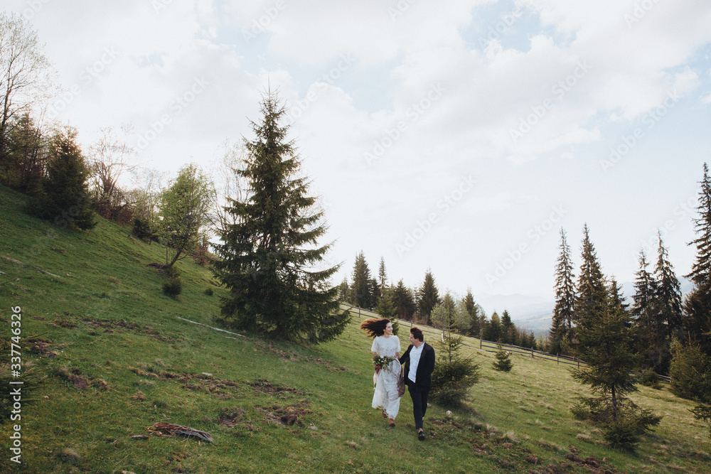 Wedding in the mountains. A young couple on a green slope among the pines, a girl in a white dress holds in her hands a bouquet of flowers and greenery with a ribbon