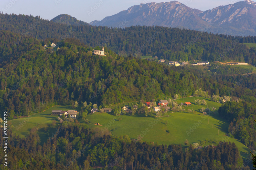 Slovenian countryside in spring with charming little village and small white church on a hill, surrounded by Julian Alps mountains, in Slovenia