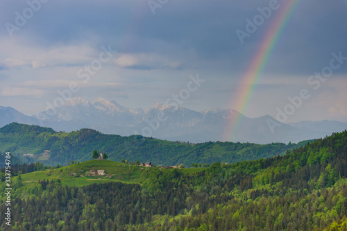 Slovenian breathtaking landscape with Julian Alps and charming little church of Sveti Tomaz (Saint Thomas) on a hill. Beautiful spring day in the mountains with rainbow after the rain, in Slovenia.