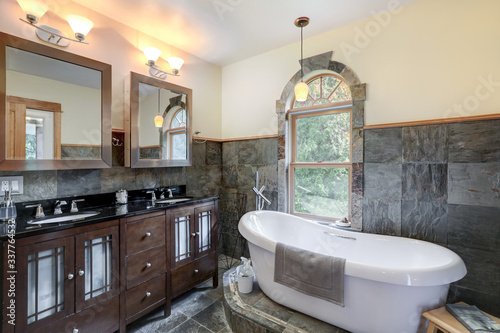 Bathroom interior with grey tiles and free standing white tub with dark wood classic vanity with black countertop and two mirrors.