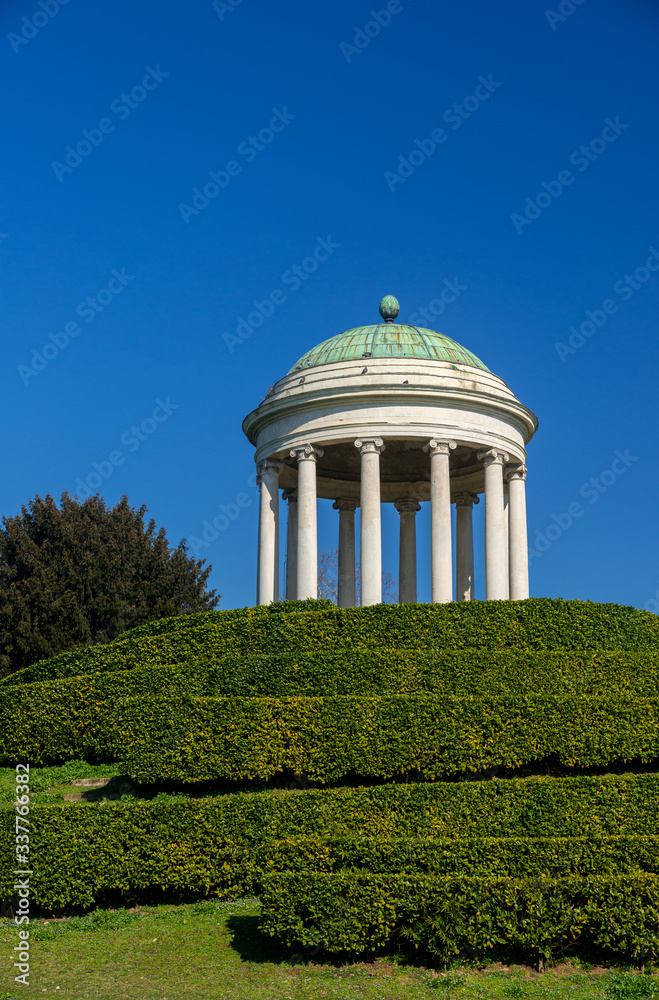A little monopteros ionic style temple stands on the hill of Parco Querini, the main public park in Vicenza. Roof in oxidized copper plates. A sunny and cloudless winter day. Boxwood bushes. Italy.
